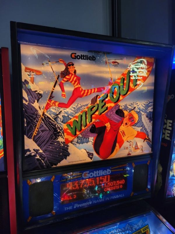 photo of title display of pinball game Wipe Out showing a blonde guy jumping with a snowboard at the front with the title "Wipe Out" on the back of the snowboard, behind him is a dark haired woman in a ski suit doing a ski jump with her left leg coming out of her crotch